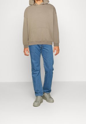 Person wearing beige hoodie, blue jeans, and gray sneakers, standing against a plain light background.