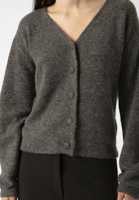 O persoană poartă un cardigan moale de culoare gri închis, cu decolteu în V și nasturi, asortat cu pantaloni negri. Accentul este pus pe textura și ajustarea îmbrăcămintei.