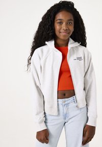 Lichtgrijze zip-up hoodie met een capuchon, voorzien van een kleine zwarte tekstprint. Draag het over een felrode crop top en lichte blauwe jeans.