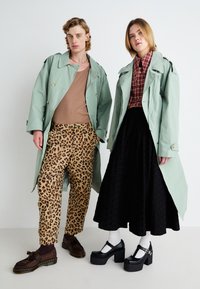 Duas modelos em trench coats verde pastel. Uma usa um top bege e calças de estampado leopardo; a outra tem uma camisa xadrez e uma saia preta texturizada.