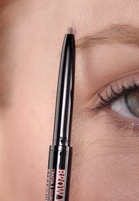 Schwarzer Augenbrauenstift, der nahe an einer Hautoberfläche gehalten wird. Der Stift hat eine feine, quadratische Spitze und ist mit der Aufschrift "Brow" in Roségold versehen. Wimpern sind sichtbar.