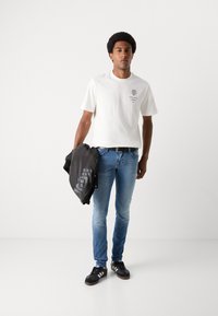 T-shirt bianca in cotone con maniche corte, stampa del logo nera, abbinata a jeans blu aderenti e scarpe da ginnastica nere, mentre tiene in mano una giacca nera.