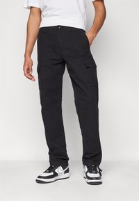 Pantalones cargo negros con bolsillos laterales, diseño de pierna recta, tejido duradero y cierre de botón. Llevados con una camiseta blanca y zapatillas.