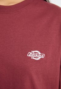T-shirt en coton bordeaux avec un logo brodé blanc sur la poitrine gauche. Encolure ronde et manches courtes avec une texture douce.