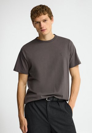 Giovane uomo con capelli corti e ricci che indossa una t-shirt grigio scuro a maniche corte infilata in pantaloni neri con cintura.