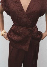 Burgundy, crinkled fabric wrap top with a deep V-neck and tie detail at the waist, paired with matching trousers. Smooth texture.
