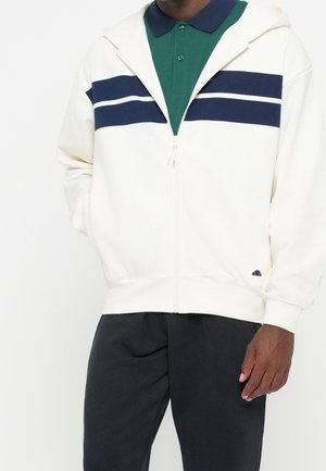 Man die een witte zip-up hoodie met marineblauwe strepen draagt over een groen poloshirt en zwarte broek, staand met één hand in de zak.