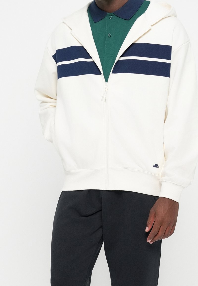 Homme portant un sweat à capuche blanc zippé avec des rayures bleu marine sur un polo vert et un pantalon noir, debout avec une main dans la poche.