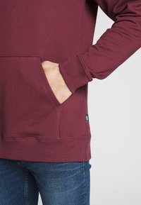 Bordový kapucňový sveter s klokanou vreckom, ribbed manžetami a dolným lemom. Mäkká textúra, viditeľné detailné prešitie a logo nášivka na dolnom ľavom okraji.