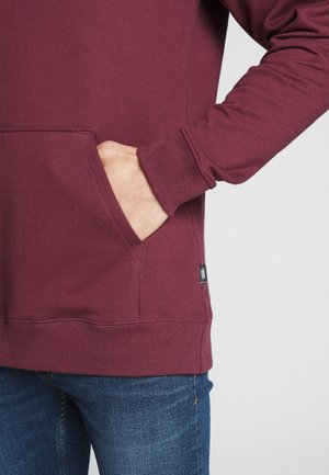 Burgunderfärgad hoodie med känguruficka, ribbade muddar och nederkant. Mjuk textur, synlig sömdekoration och logotag på nedre vänstra sidan.