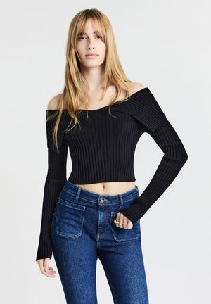 Schwarzer, cropped, schulterfreier, gerippter Pullover mit langen Ärmeln. Kombiniert mit hochtaillierten blauen Jeans mit vorderen Taschen.