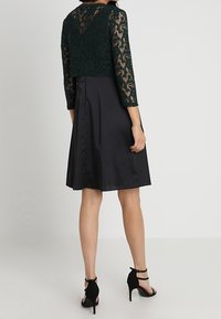 Žena v černé sukní ke kolenům a tmavě zeleném krajkovém topu s dlouhými rukávy, stojící zády ke kameře.