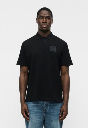 Bărbat purtând un tricou polo negru cu mâneci scurte și un logo subtil pe piept, și blugi albaștri, pe un fundal simplu.