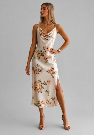 Femme portant une robe fleurie crème sans manches avec une fente latérale, associée à des sandales à talons nude et des bijoux minimalistes sur un fond gris.