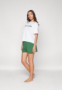 T-shirt blanc à manches courtes avec le logo "LACOSTE" en vert, associé à un short vert. Tissu en coton, coupe décontractée, texture lisse, design simple.