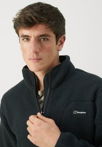 Giacca in pile scura con colletto alto e zip, realizzata in un tessuto testurizzato e morbido. Logo Berghaus sul lato sinistro superiore.