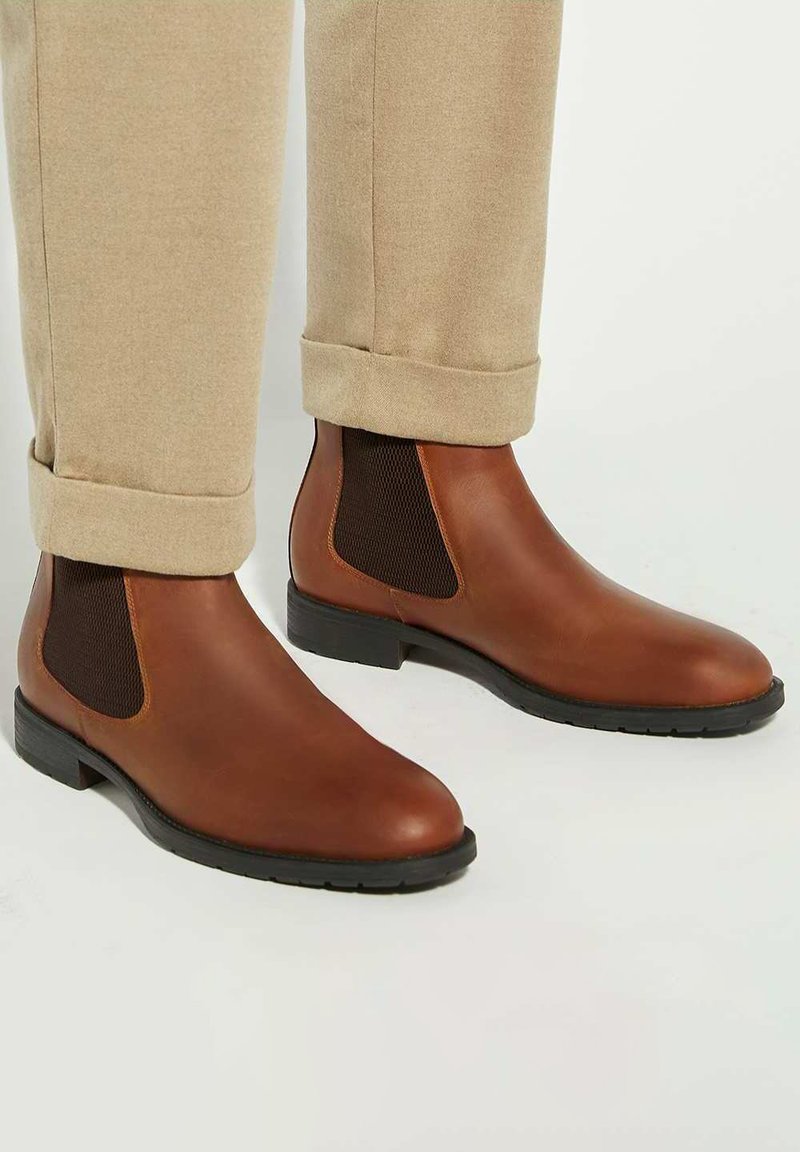 Brown leather Chelsea boots with fabric side panels, rounded toe, and black rubber sole. Paired with rolled beige pants.