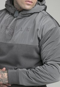Felpa grigia con cappuccio, sezione inferiore in morbido pile, parte superiore liscia, cappuccio con cordini e logo ricamato sul petto.
