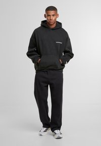 Giovane uomo in piedi con le mani in tasca, indossando un hoodie nero, pantaloni neri e sneaker bianche, su uno sfondo grigio chiaro uniforme.