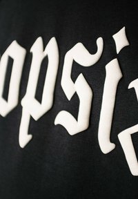 Svart stoff med hvite, hevede gotiske bokstaver med buede aksenter og skarpe kanter. Teksturen ser myk og plysjaktig ut.
