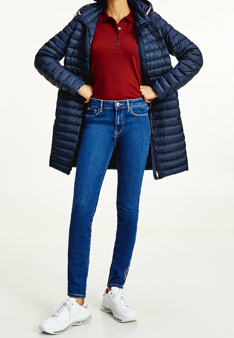 Personne portant un manteau matelassé bleu marine sur un polo bordeaux, un jean skinny bleu et des baskets blanches, debout les mains sur les hanches.