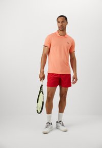 Polo skjorte i lys oransje med en svart logo, kombinert med røde shorts. Holder en tenniskølle. Hvite sokker og sportssko.