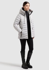 Prošívaná bunda ve světle šedé barvě s kapucí, prošívaným vzorem, černými zipy a dvěma předními kapsami. Nošena přes černé legíny a boty.