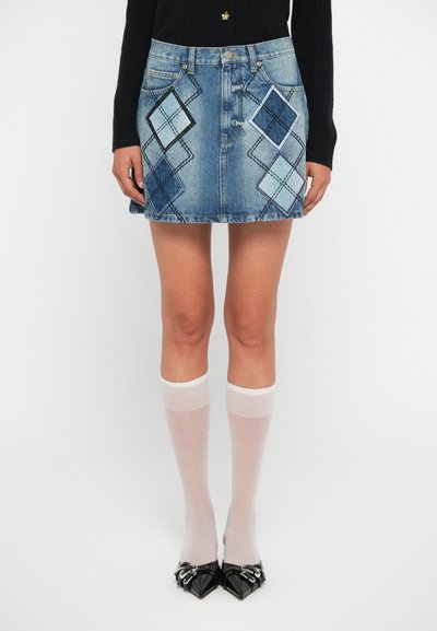 Jupe en denim mini avec des motifs de diamants dans différentes nuances de bleu, poches avant et ourlet effiloché. Associée à des chaussettes blanches jusqu'aux genoux.
