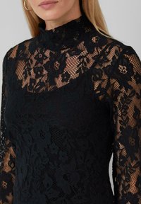 Haut en dentelle noire avec un col haut, des motifs floraux et des manches longues avec des détails transparents. La texture du tissu est délicate et complexe.