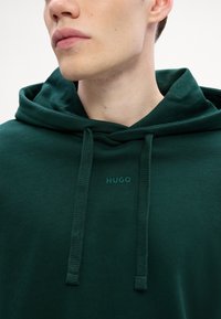 Tamsiai žalias džemperis su kapučiu, pagamintas iš minkštos medžiagos, su raišteliu, kengūros kišene ir atitinkamos spalvos "HUGO" logotipu ant krūtinės.