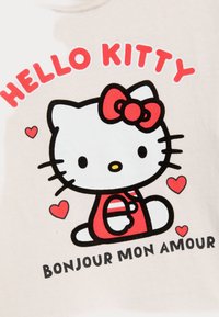 Beige t-shirt met een cartoonkat met een rode strik, rode accenten en een zwarte omtrek. De tekst luidt "HELLO KITTY" en "BONJOUR MON AMOUR."