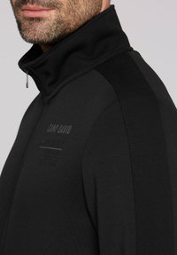 Zwarte zip-up jacket met een hoge kraag, voorzien van gestructureerde schouderpanelen, reliëf logo detail en een slanke, getailleerde snit.