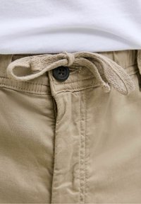 Beiget puuvillahousut, joissa on kiristysnaru ja napitettava sulkeminen. Housuissa on sileä pinta ja näkyvät ompelusaumat vyötärön läheisyydessä.