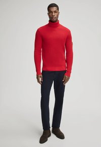 Rode gebreide coltrui met geribbelde textuur, gedragen met donkerblauwe slim-fit broek en bruine enkellaarsjes.