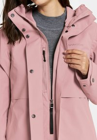 Vaaleanpunainen vedenpitävä takki mustalla vetoketjulla ja yksityiskohdilla, jossa on huppu, nepparit ja harmaa neule sen alla.