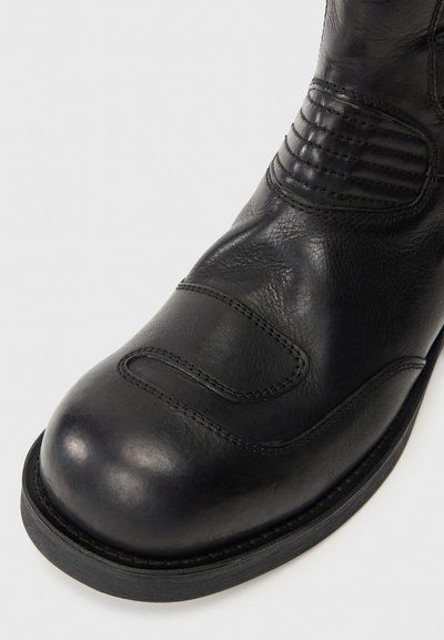 Boot de piele neagră cu vârf rotunjit, superior texturat cu accente striate și o talpă robustă din cauciuc. Cusături vizibile pe margine.