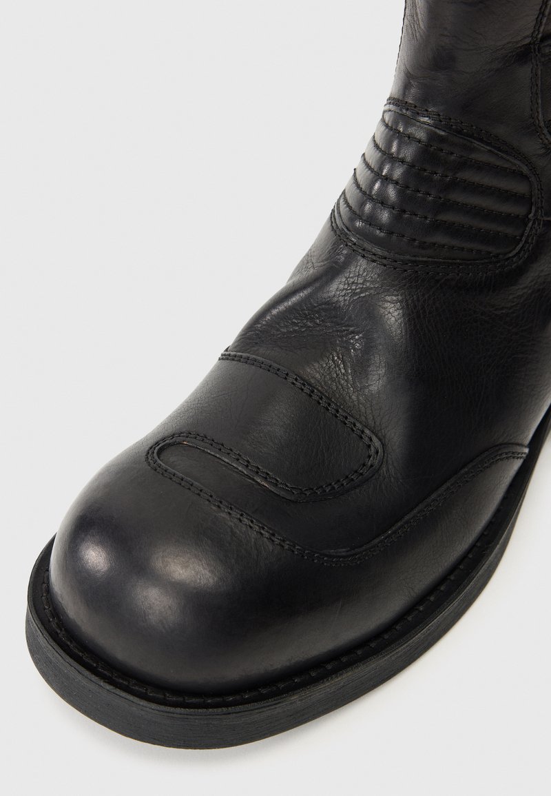 Botte en cuir noir avec un bout arrondi, tige texturée avec des accents nervurés et une semelle en caoutchouc robuste. Couture visible le long du bord.