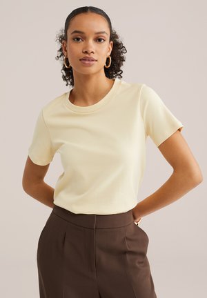 Frau mit lockigem Haar, die goldene Creolen, ein cremefarbenes kurzärmeliges Oberteil und eine braune Hose mit hohem Bund trägt, steht mit den Händen hinter dem Rücken.