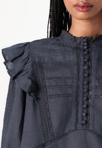 Dark blue blouse featuring ruffled sleeves, intricate lace accents, button closure, and textured stripes across the bodice.