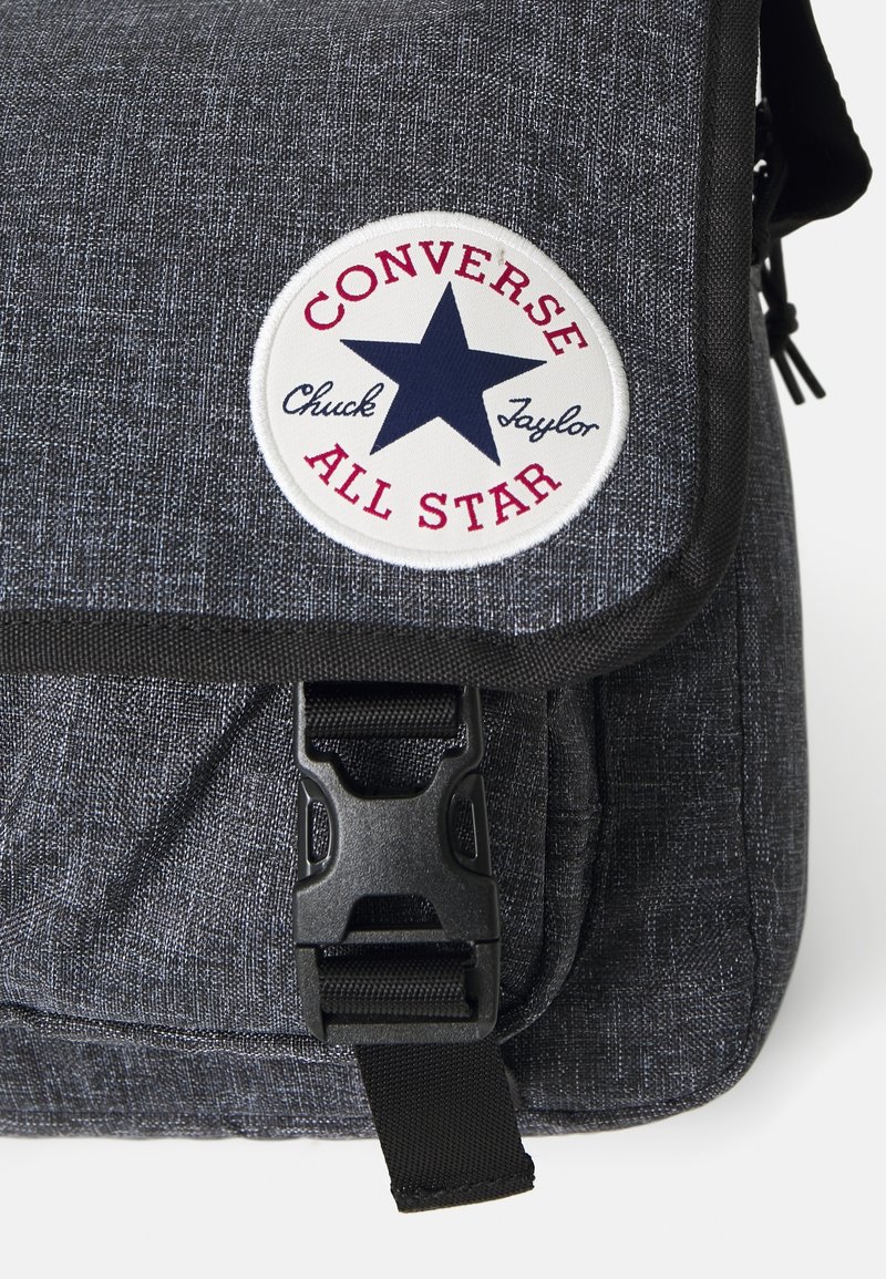 Bolso mensajero de tela gris con una textura suave, que presenta un parche redondo con la inscripción "Converse All Star" y un diseño de estrella, además de un cierre de hebilla de plástico.