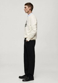 Bela majica s kapuco z grafičnim tiskom, v kombinaciji s temnimi hlačami in črnimi čevlji. Tkanina se zdi gladka, z sproščenim krojem.