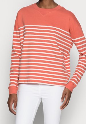 Persona vistiendo una camisa de manga larga de rayas horizontales coral y blancas con pantalones blancos, de pie contra un fondo liso.