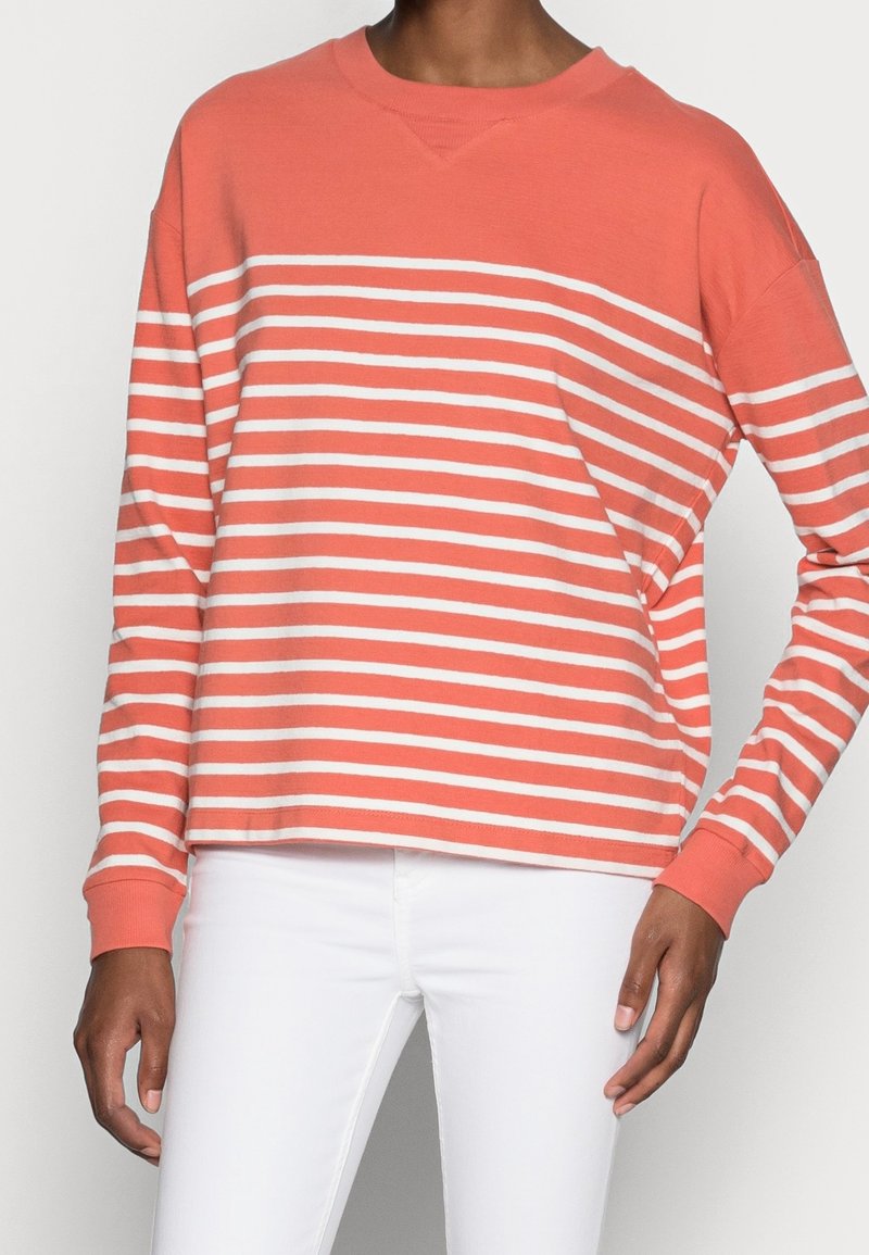 Personne portant une chemise à manches longues rayée horizontalement corail et blanche avec un pantalon blanc, debout devant un fond uni.