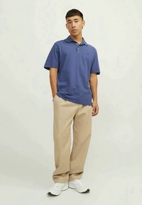 Muškarac stoji s rukama u džepovima, odjeven u plavu polo majicu, bež hlače i bijele tenisice, nasuprot jednostavnoj svijetloj pozadini.