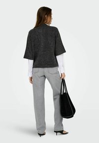Femme portant un pull à manches courtes gris foncé sur une chemise blanche, un jean gris clair, des sandales noires à talons, tenant un grand tote noir, de dos.