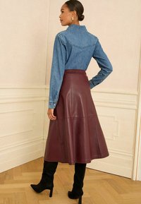 Denimhemd mit spitzem Kragen, kombiniert mit einem midi Rock aus burgunderfarbenem Kunstleder. Schwarze Absatzstiefel runden den Look ab.