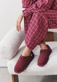 Scarpe slip-on color bordeaux in tessuto morbido con punta arrotondata, abbinate a pantaloni da pigiama a quadri rossi e blu, poggiate su una sedia bianca con texture.