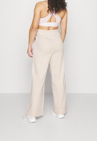 Pantalones deportivos beige de pierna ancha con textura suave, con rayas blancas en los costados y cintura elástica. Combinados con un sujetador deportivo rosa pálido.