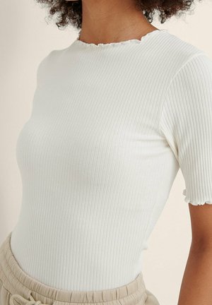 Woman wearing a white ribbed short-sleeve top with ruffled edges and beige elastic waist pants, shown from neck to waist.