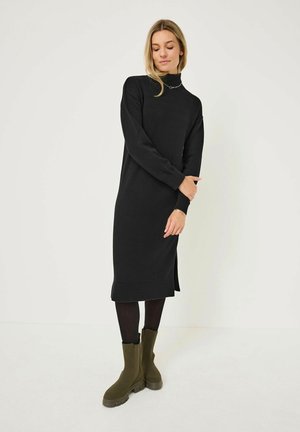 Robe en tricot noire avec un col montant, des poignets côtelés et des fentes latérales ; associée à des bottines vertes à bout carré avec des semelles épaisses et une finition mate.
